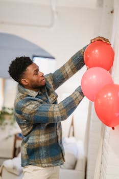 Side view of young bearded African American male in casual clothes standing near white wall and decorating room with inflatable balloons