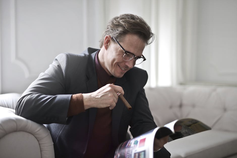 Cigar Room Smiling rich gentleman wearing classy suit and stylish eyeglasses sitting with cigar on leather sofa in luxury living room and reading magazine