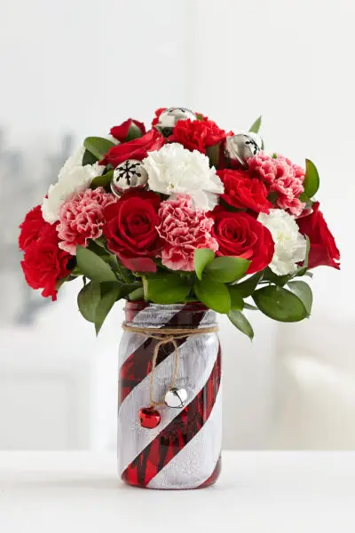Finished red and white painted glass mason jar centerpiece vase with flower bouquet of red roses, pink and white carnations and jingle bells