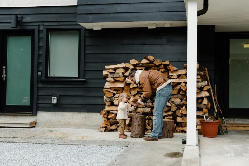 Father and toddler daughter stacking firewood outside modern home.