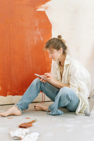 Adult woman taking a break from painting walls, sitting on floor, using smartphone for relaxation.