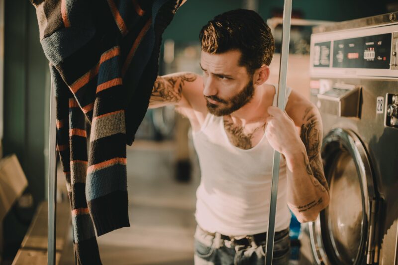 Bearded man with tattoos using dryer in laundromat wearing tank top.