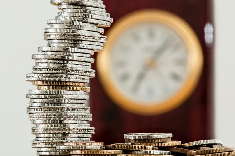 A close-up image of stacked coins with a blurred clock, symbolizing time and money relationship.