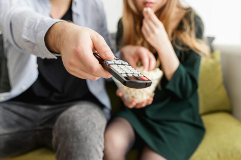 A couple enjoying a relaxing movie night, sitting on a sofa with popcorn and a TV remote.