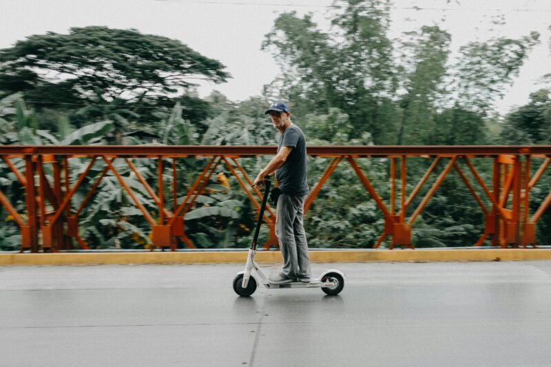 Adult man rides an electric scooter across a bridge surrounded by greenery.