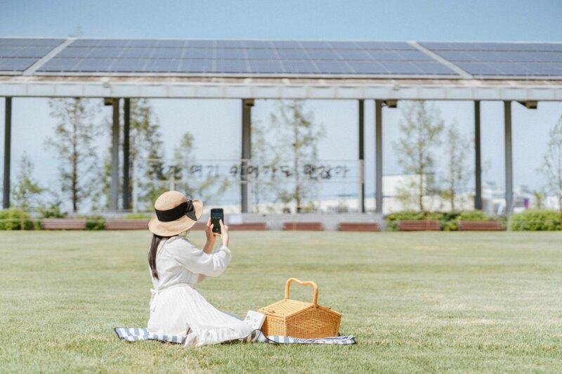 A woman in a white dress and sun hat takes photos during a picnic in a sunny park. Relaxing outdoor scene.