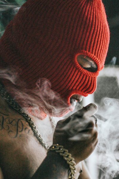 A person wearing a red balaclava smoking, with visible tattoos and gold chain, in an urban environment.