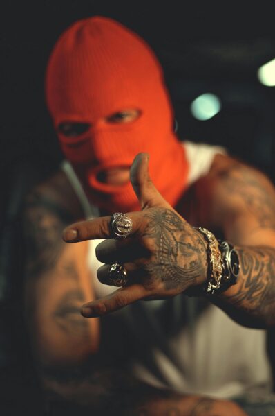 A tattooed man wearing a red ski mask gestures in a dimly lit urban studio.