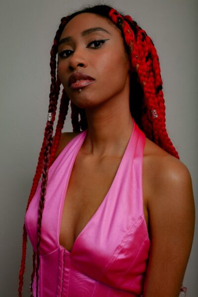 A young woman poses fashionably in a pink dress with striking red braided hair indoors.