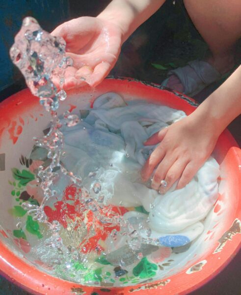 Close-up of hands washing clothes in a red basin outdoors. Sunny day.