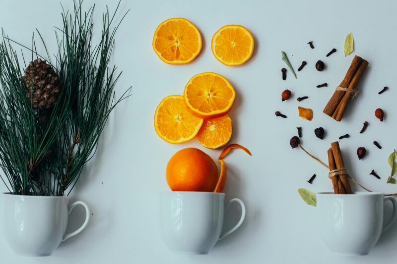 Flat lay of pine branches, citrus slices, and spices in cups on white background.