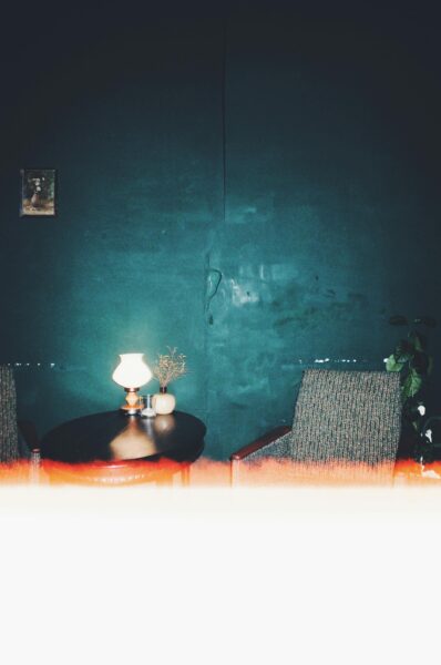 A vintage room interior featuring armchairs, a lamp, and a vase against a dark teal wall with film-like imperfections.