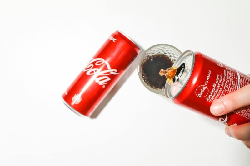 Close-up of cola being poured from a red can into a glass against a white background.