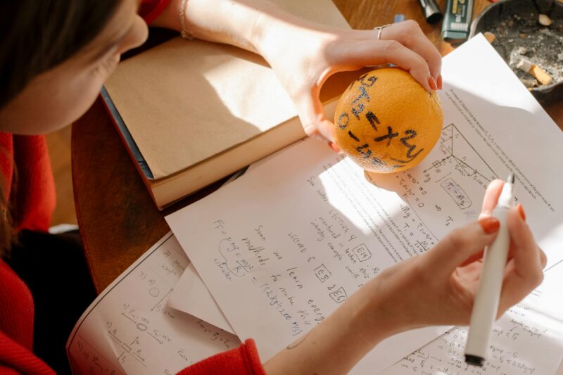 Student studying math creatively using an orange and notes in warm indoor light.