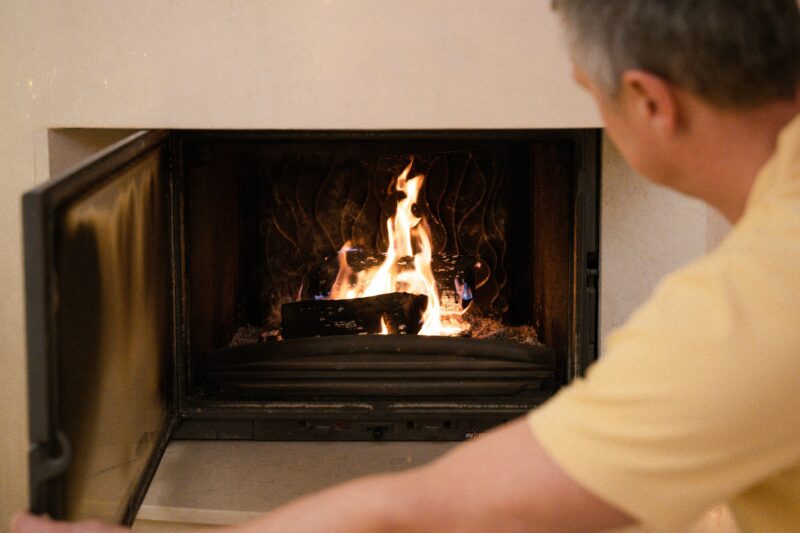 A man in a yellow shirt tends to the burning fire in a cozy indoor fireplace.