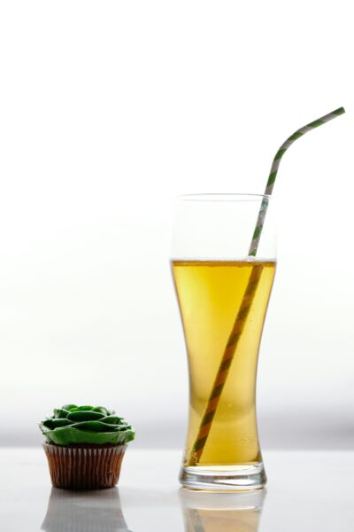 Close-up of a green-frosted cupcake next to a beer glass with a straw, white background.