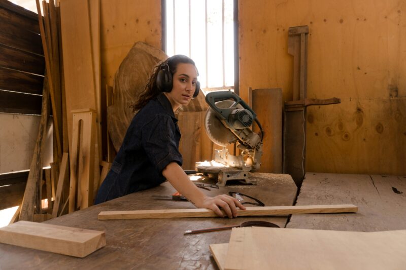 Caucasian woman works with a miter saw in a carpentry workshop, focused on precise wood cutting.