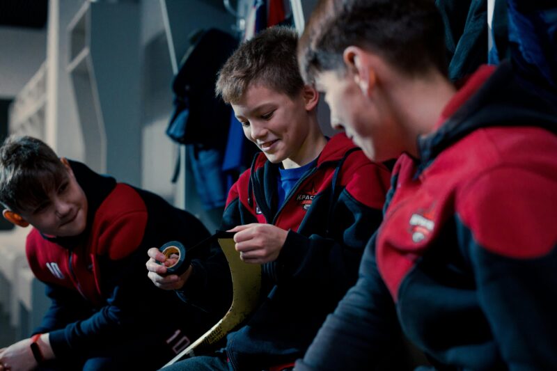 Three young hockey players in a dressing room, preparing equipment for a game.