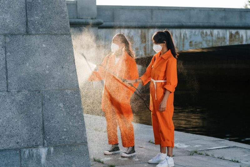 Two people in orange jumpsuits performing outdoor cleaning while wearing face masks.