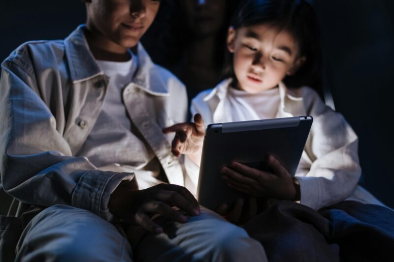 Siblings sitting in a dimly lit room, using a tablet for entertainment or education.