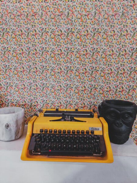 Vintage yellow typewriter on a table with a colorful floral wallpaper background.