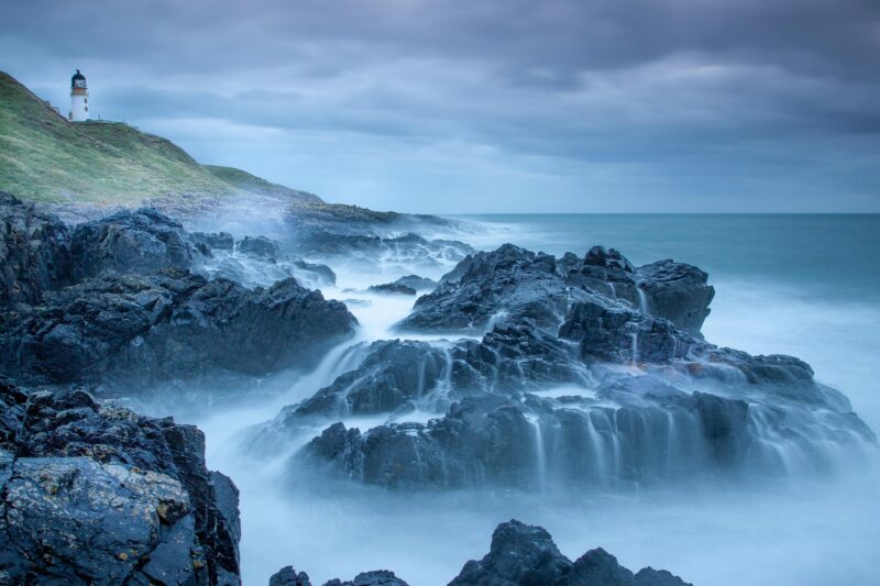 Captivating seascape of a lighthouse and rugged coastline with waves crashing against the rocks in Scotland.