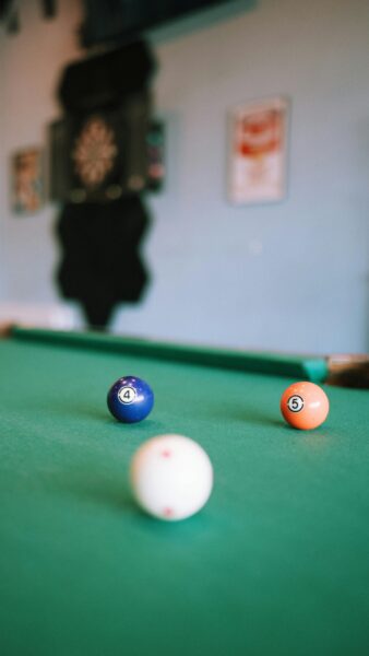 Vibrant billiard balls on a green pool table with dartboard in the background.