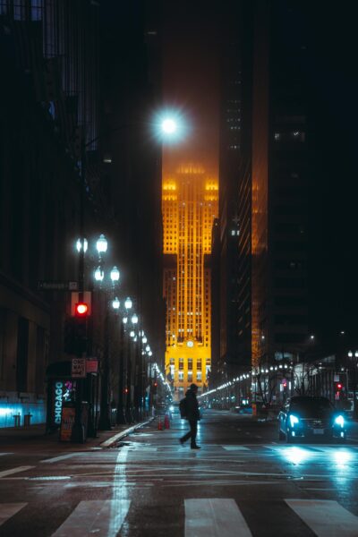 A lone figure crosses a city street at night, illuminated by a glowing skyscraper.