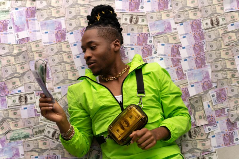 An African American man in a bright green jacket surrounded by various currencies, showcasing wealth and style.