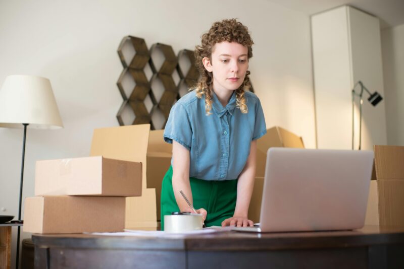 A young woman working on her laptop surrounded by cardboard boxes, indicating online business operations.