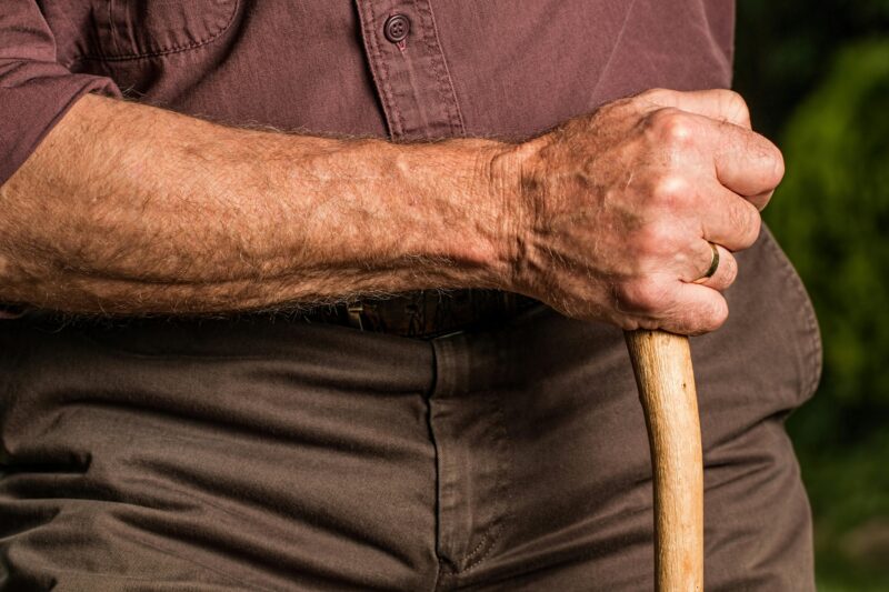 Close-up of a senior adult's hand gripping a wooden walking cane outdoors, symbolizing support and aging.