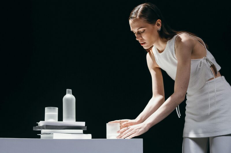 A young woman in a white dress arranging monochrome vases on a table with a black background.