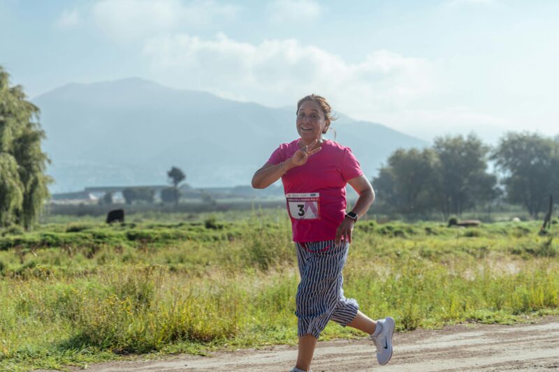 A joyful woman jogs on a rural dirt road, enjoying a sunny day with scenic mountain views.