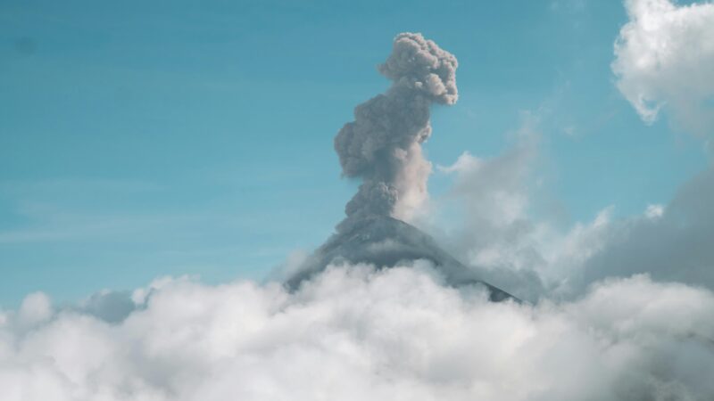 Dramatic view of Acatenango Volcano erupting amidst clouds under a clear blue sky.