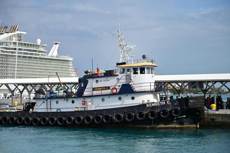 A tugboat named Kenneth C docked at a busy port next to a cruise ship on a clear day.