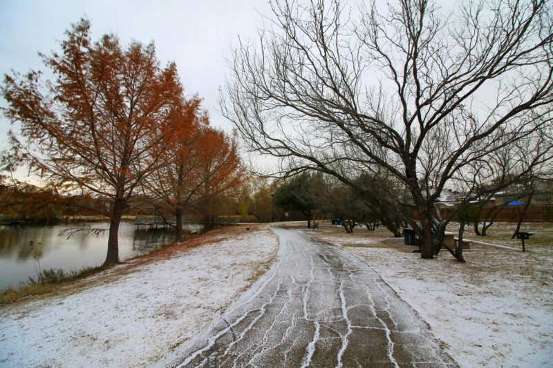 A tranquil winter scene in San Antonio, Texas, showcasing a snow-covered path lined with trees beside a serene lake.