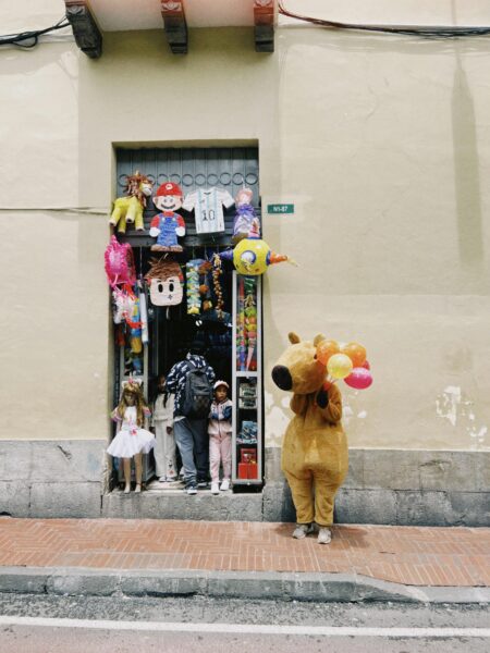 A lively toy store with piñatas and a bear costume holding balloons outside.