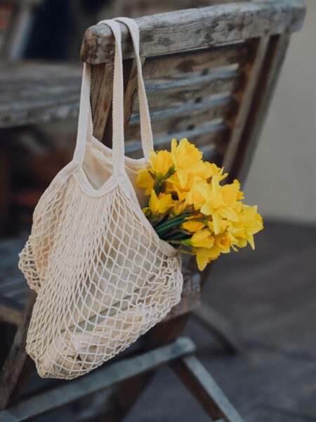 Mesh bag with bright yellow daffodils hanging on a rustic wooden chair outdoors.