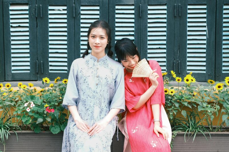 Two women in traditional attire sitting outdoors with flowers and a shuttered window backdrop.