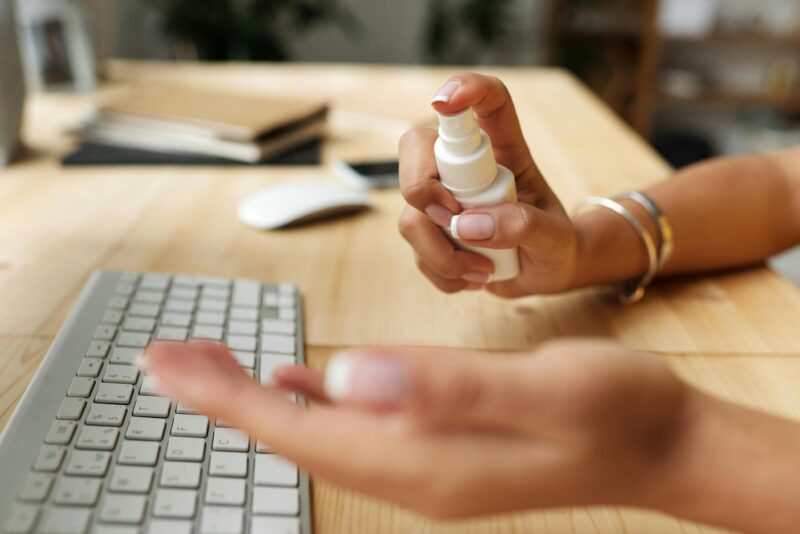 Person sanitizing hands at a desk with spray bottle, keyboard, and computer mouse.