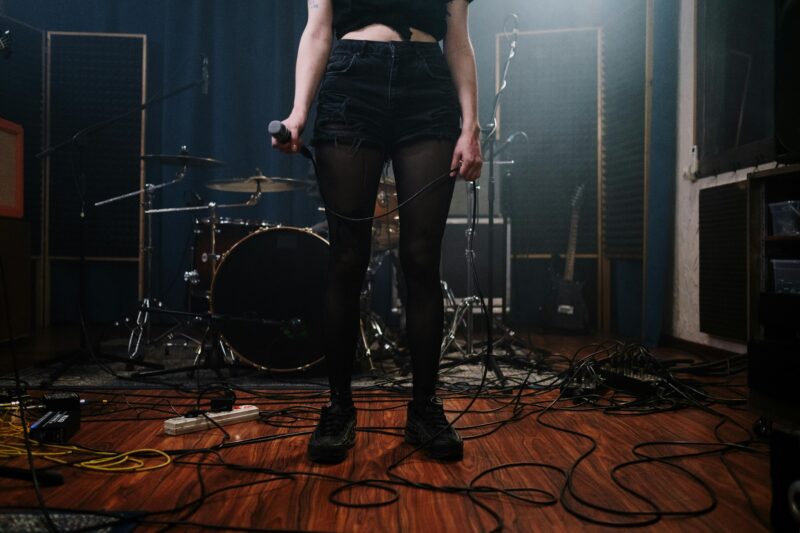 A woman in denim shorts holds a microphone on a wooden floor, surrounded by band equipment.