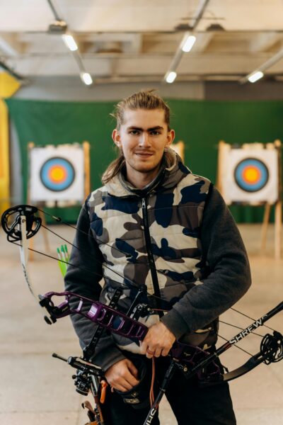 Portrait of a confident male archer holding a compound bow indoors with archery targets in the background.