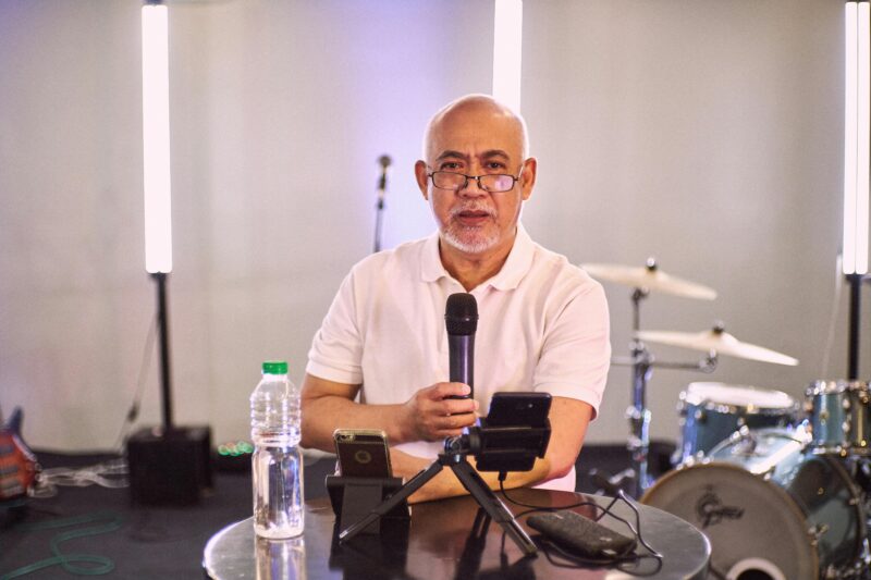 Senior man in a studio setting using a microphone with a drum set in the background.