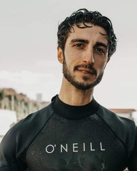 Pensive man in wetsuit with wet hair posing outdoors with city background.