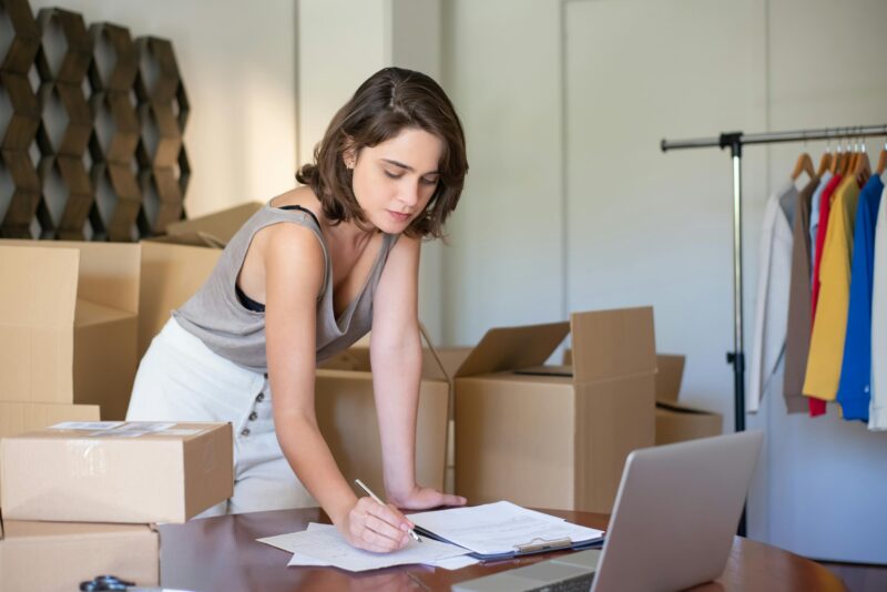 Young woman managing her online clothing business from home office with boxes and laptop.