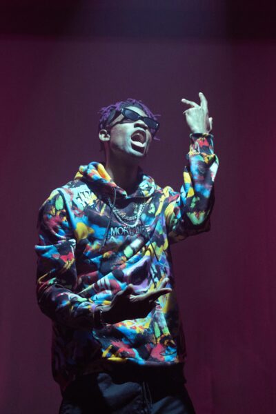 A rapper passionately performing on stage in vibrant streetwear and sunglasses under dramatic lighting.