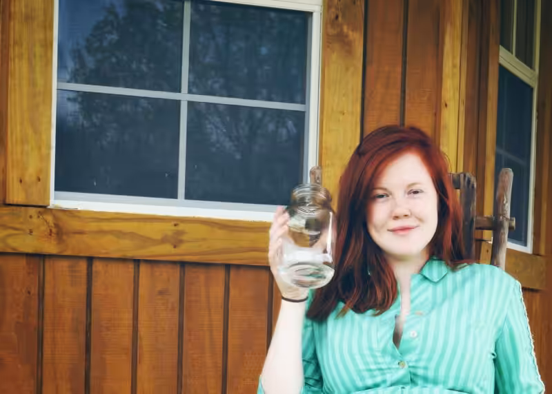 Smiling young woman holding a glass jar on a wooden porch in the countryside.