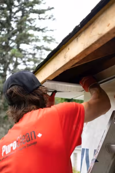 A construction worker in a red shirt installs a gutter under a wooden roof, focusing on precision.