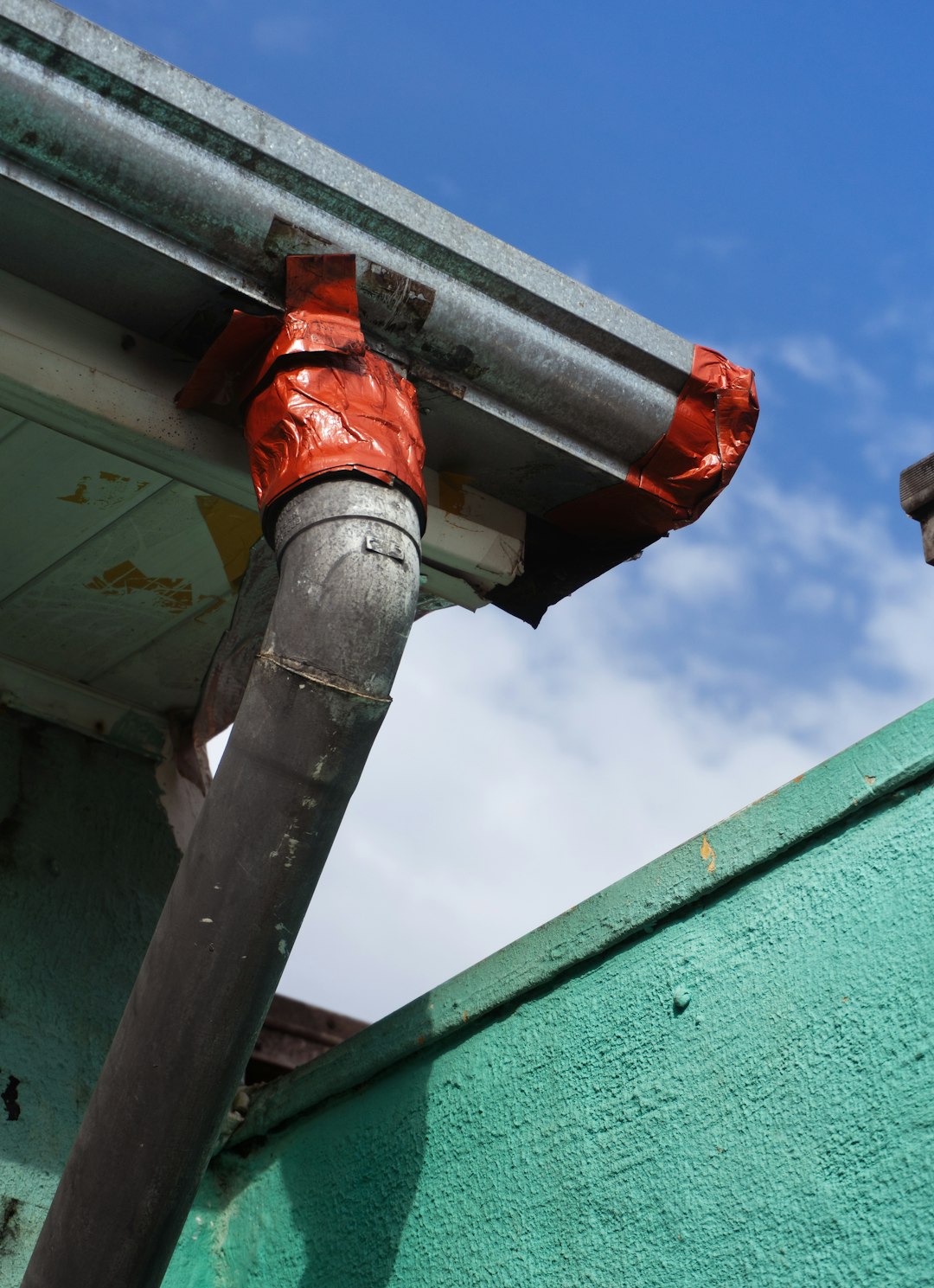 A rusted drainpipe held together with orange tape on a weathered teal wall.