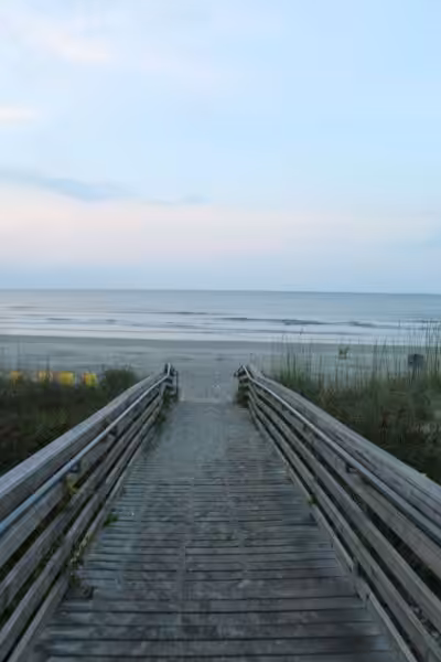 A tranquil beach scene with a wooden pathway leading to the ocean under a clear sky.